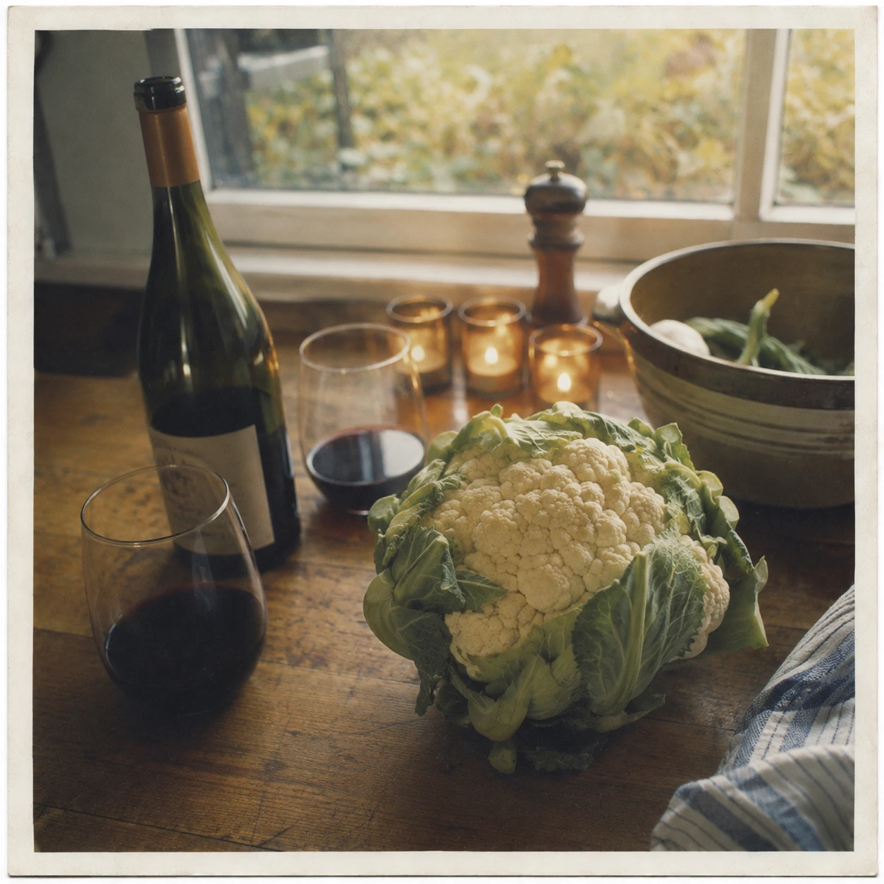 A cauliflower on a kitchen counter beside an open bottle of wine and lit candles, suggesting a quiet dinner at home.