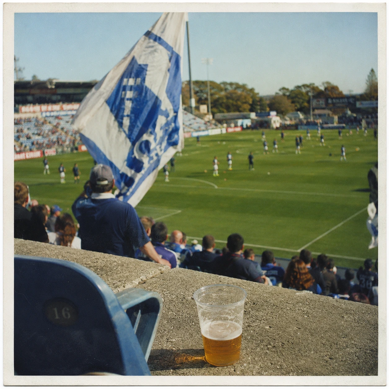 View from the stands at the Silverdale Sirens women's cup final, showing a supporter with a large flag, plastic stadium seats, a cup of beer, and players warming up on the pitch.